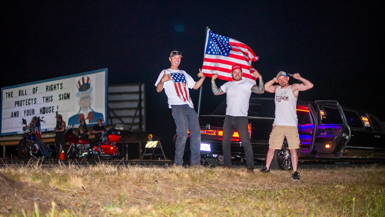 From left to right, Drew Webb, Cameron Bluhm, and Austin Jones stand outside a limousine while an American flag waves behind as they "protect" the Hamilton sign from Antifa threats Tuesday night south of Chehalis.