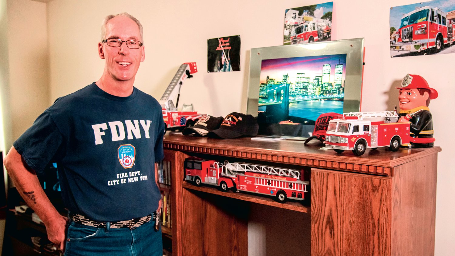 Charles Tippett Jr. smiles and poses for a photo next to a collection of fire hats and engines displayed next to an illuminated photo of the New York City skyline in his Centralia residence. Tippett Jr. sports an “FDNY” shirt he received while working as a firefighter in the city, where he helped victims during the events that unfolded on Sept. 11, 2001. "That's my boys right there," he said Saturday while describing the fire trucks displayed in his living room.