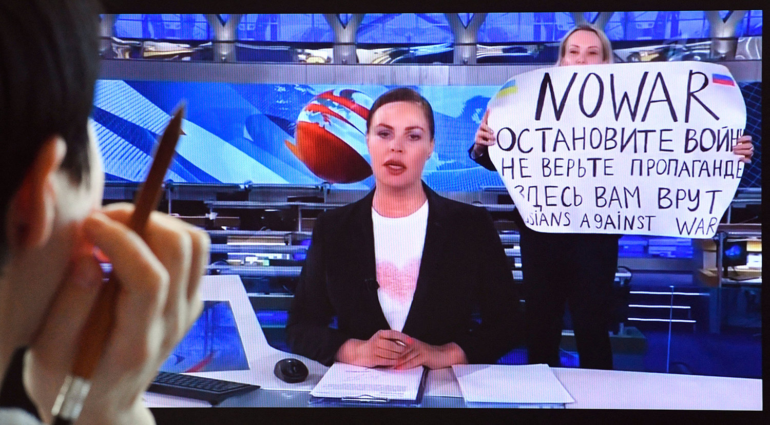 As news anchor Yekaterina Andreyeva, center, discusses Russia's relations with Belarus, Marina Ovsyannikova, right, burst into view, holding up a handwritten poster saying "No war" in English. (AFP via Getty Images/TNS)