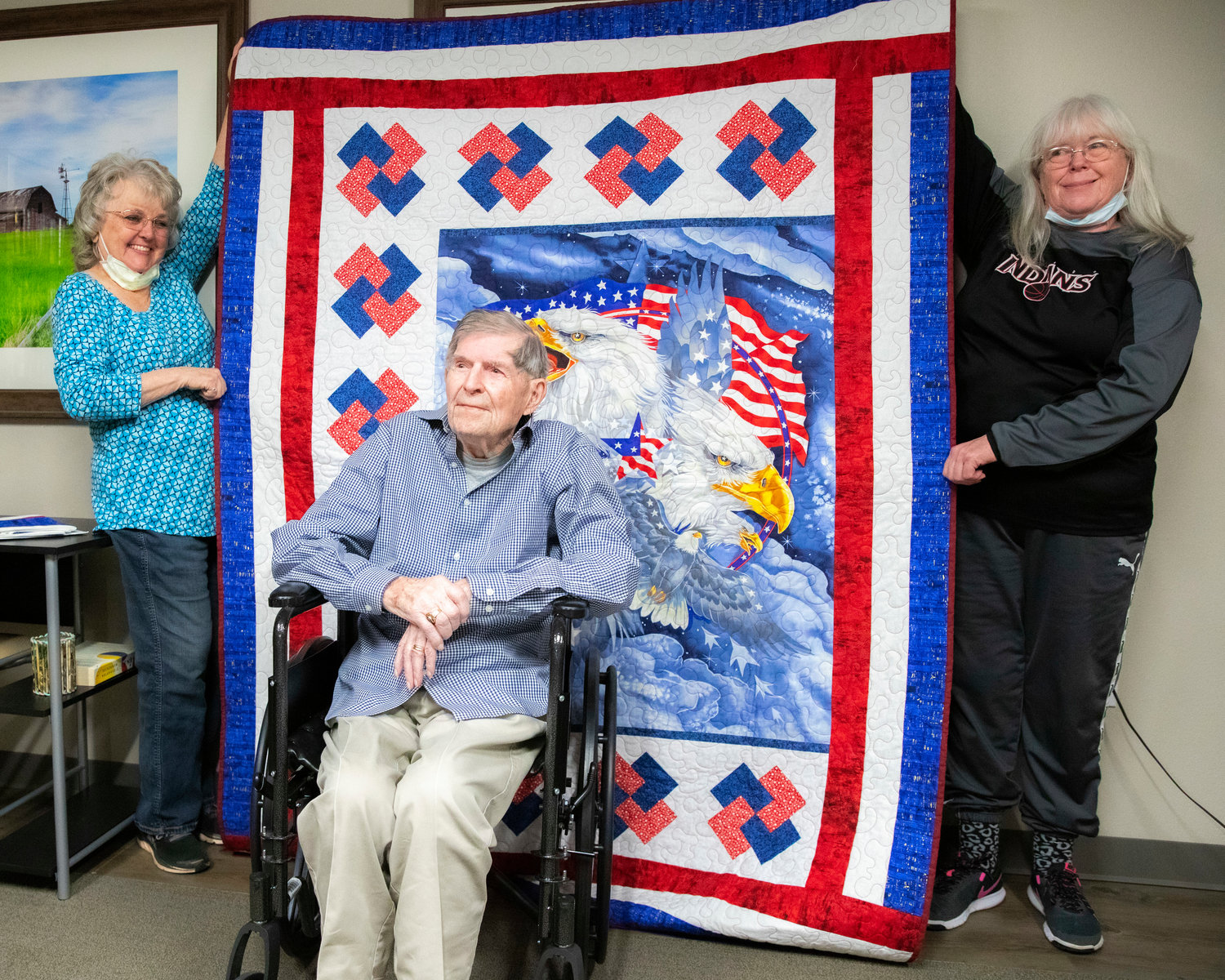 Debbie Austons and Connie Carter smile and hold up a blanket for James F. Van Ackeren, a WWII veteran who served as a staff sergeant in the U.S. Army, during a Quilts of Valor ceremony at Woodland Estates in Chehalis on Thursday.