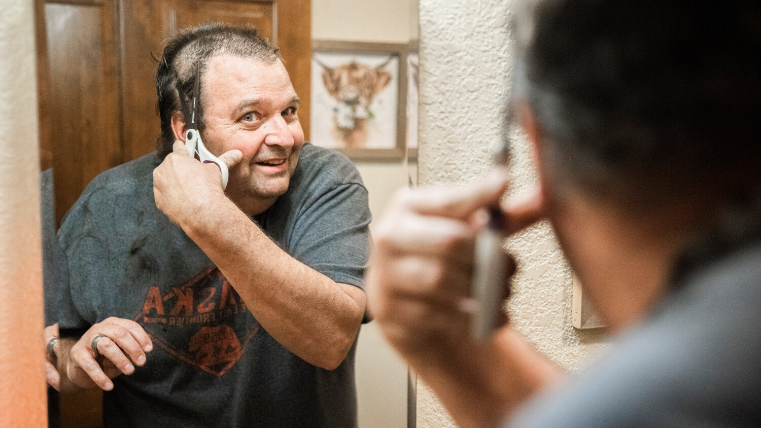 Ken Wiseman smiles into the mirror while holding a pair of scissors to his hair making cuts to raise awareness for Tonja Vlach, a receptionist at I-5 Toyota, who is undergoing chemotherapy for breast cancer and an auction the family is holding online to raise funds this weekend.