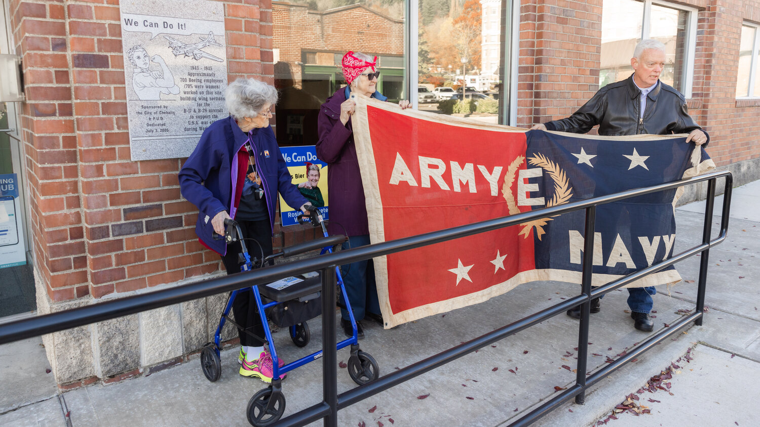Peter Lahmann, far right, talks about the history behind a flag displayed in front of a “Rosie The Riveter” plaque outside the Lewis County Public Utility building Thursday afternoon in Chehalis. The Army-Navy “Excellence in Production” flag was created in 1942 to recognize contributions made to help war efforts.